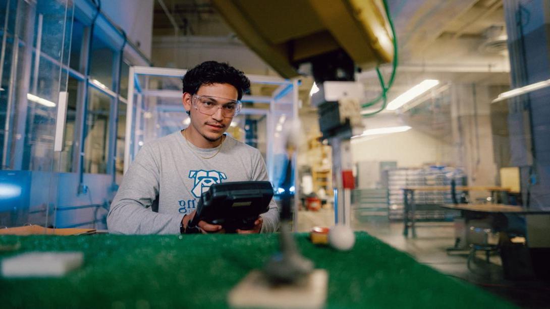 A Kettering industrial engineering student wears safety goggles and controls and industrial robot arm in the Polymer Lab